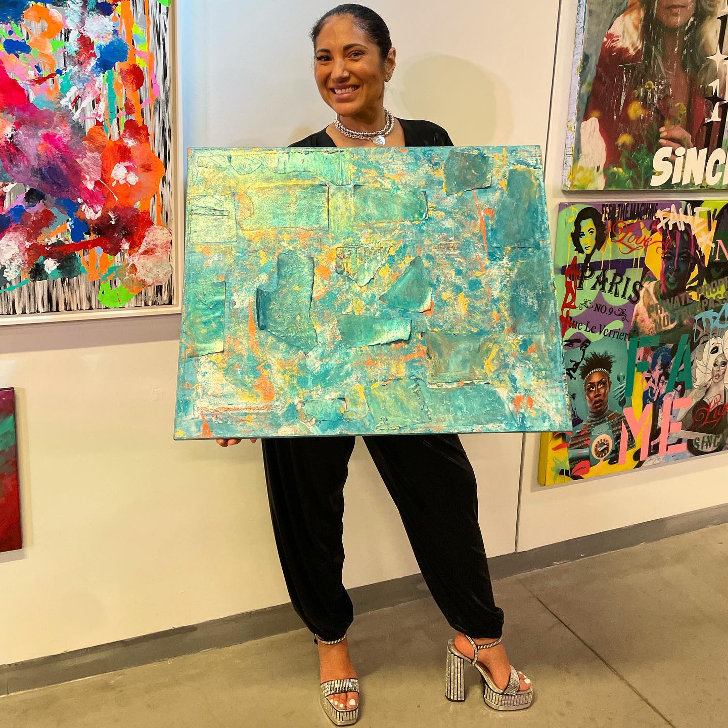 A collector holds a teal painting in a gallery surrounded by other works.