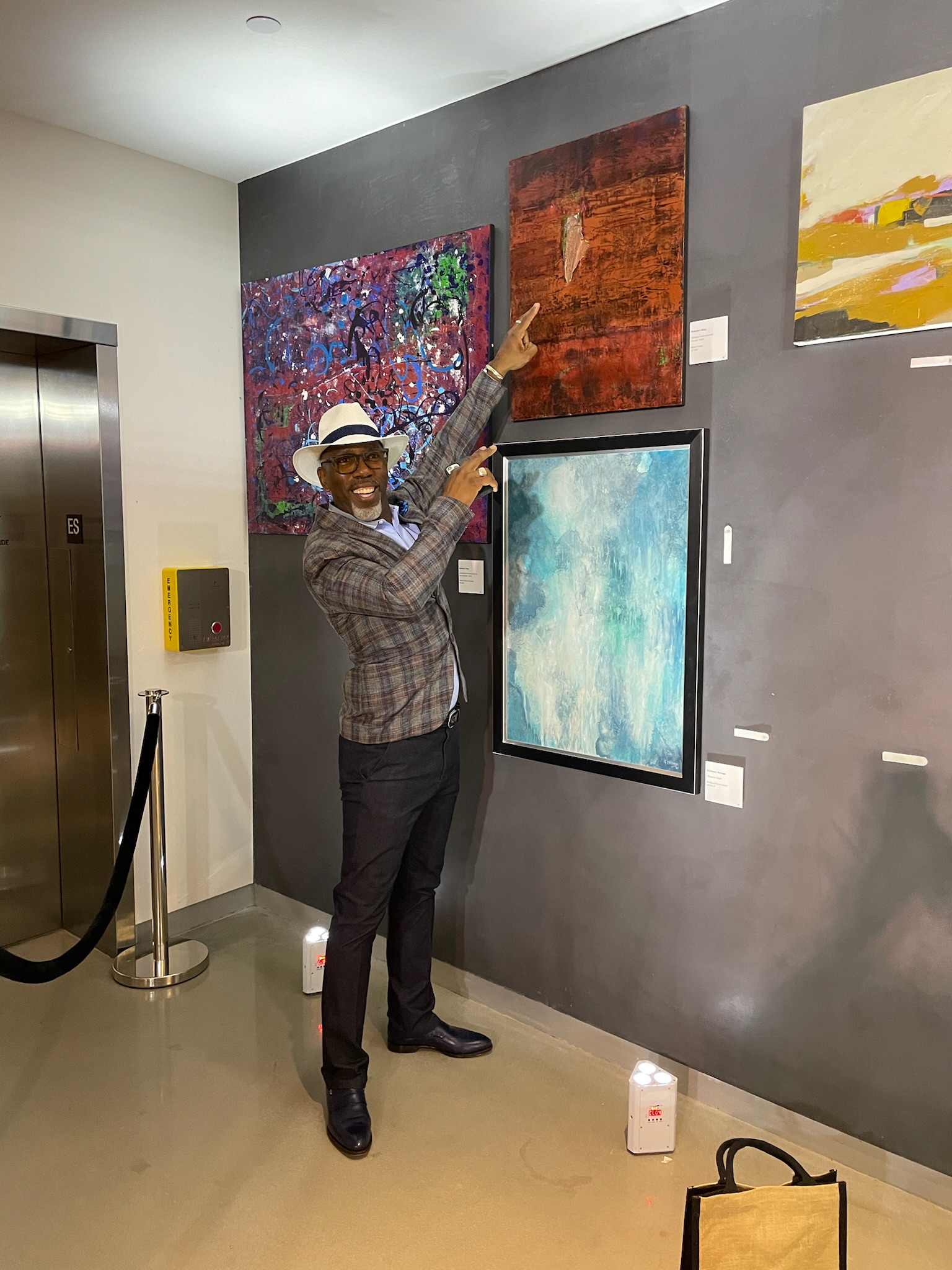 A visitor in a panama hat points to a canvas on a dark gallery wall.